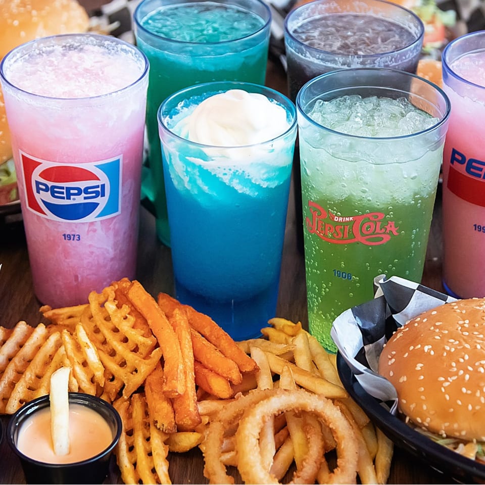 Kirt's Drive-In burgers, fries, onion rings, and colorful Pepsi drinks on a wooden table.