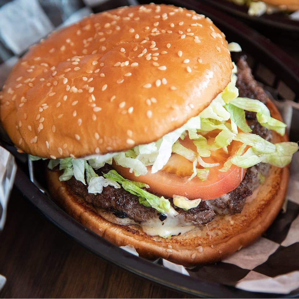 A Kirt's Drive-In burger with lettuce, tomato, and melted cheese on a sesame seed bun.