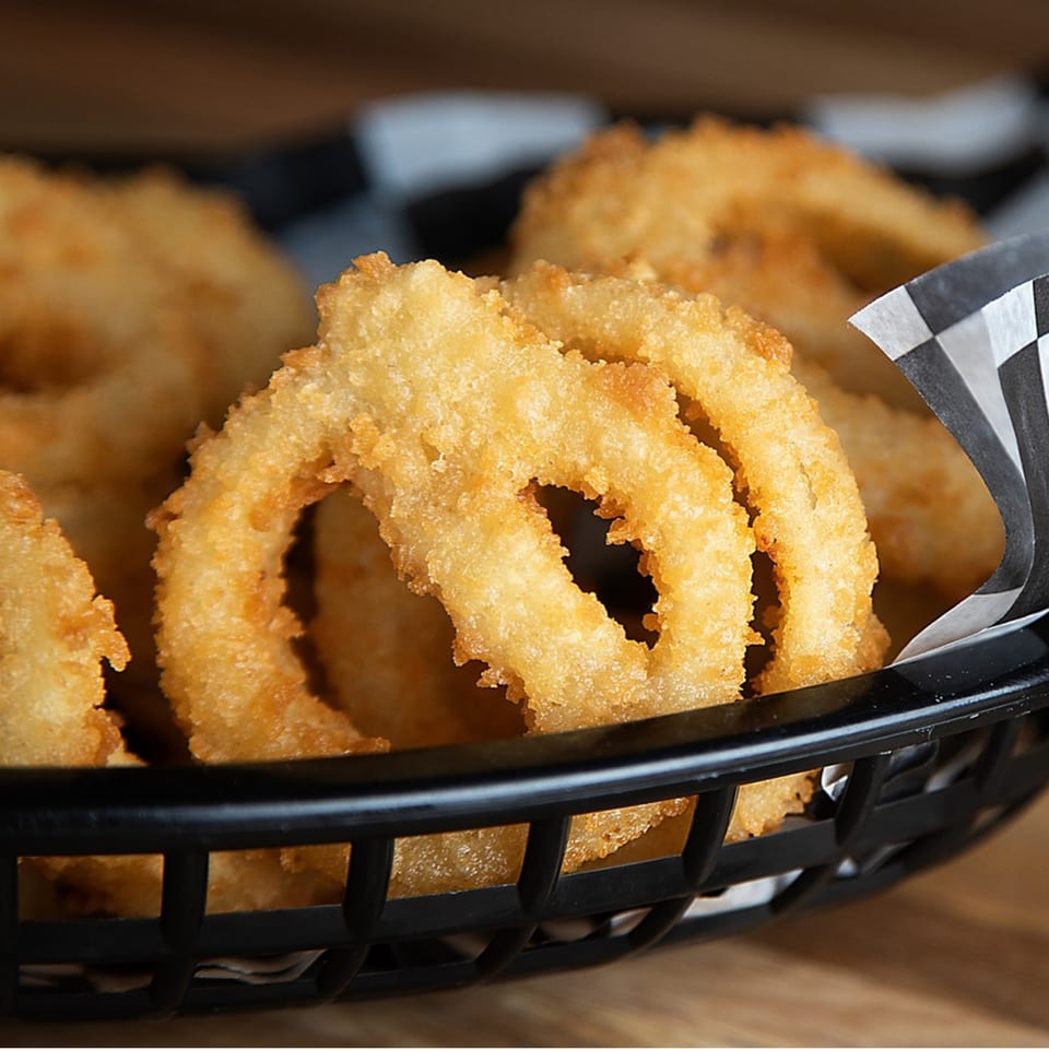 Golden-brown, crispy onion rings in a black basket with checkered paper from Kirt's Drive-In.