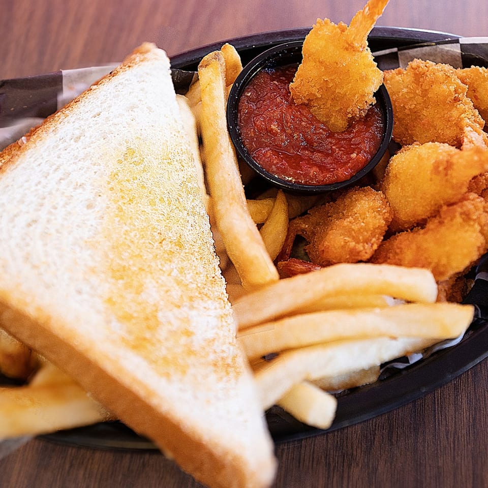 A basket of golden-brown fried shrimp, french fries, and toasted bread, with one shrimp dipping into red sauce.